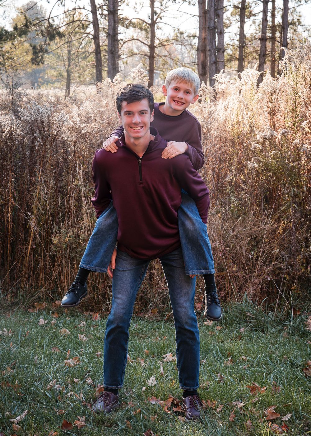 Teen boy giving younger blonde boy a piggyback ride outdoors in autumn field with golden grasses.