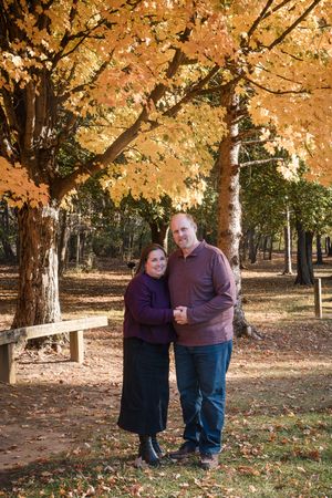 Couple embracing under vibrant orange autumn maple trees in a park, wearing purple and mauve sweaters, smiling warmly.