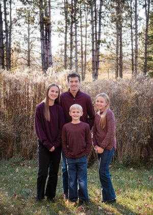 Four siblings pose outdoors in coordinating burgundy outfits among tall grasses and pine trees in a fall family portrait.