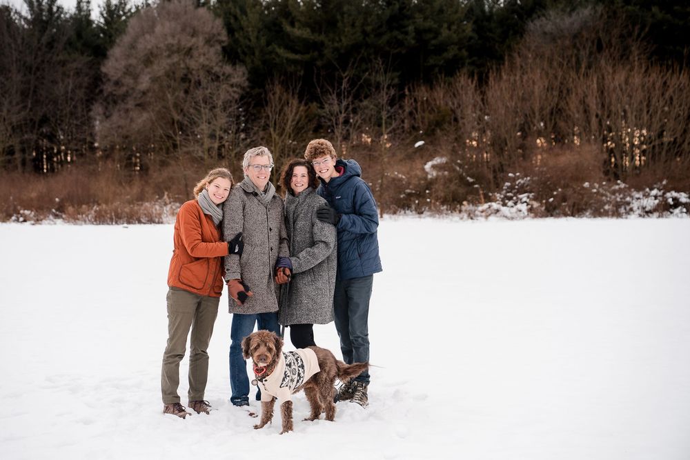 Family of four posing with their dog in a snowy winter landscape surrounded by bare trees.