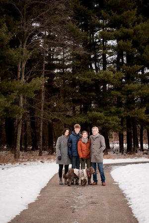 Family of four with dogs posing on a snowy path surrounded by tall pine trees in winter.