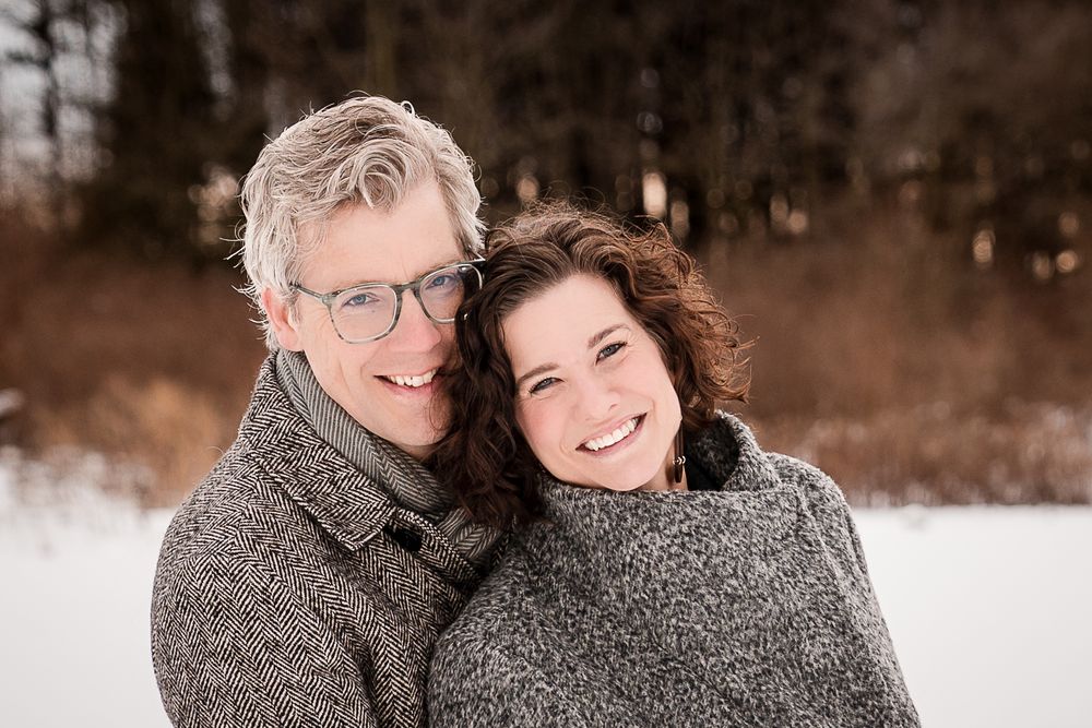 Smiling couple in gray coats posing together in a snowy winter outdoor setting with trees in the background.