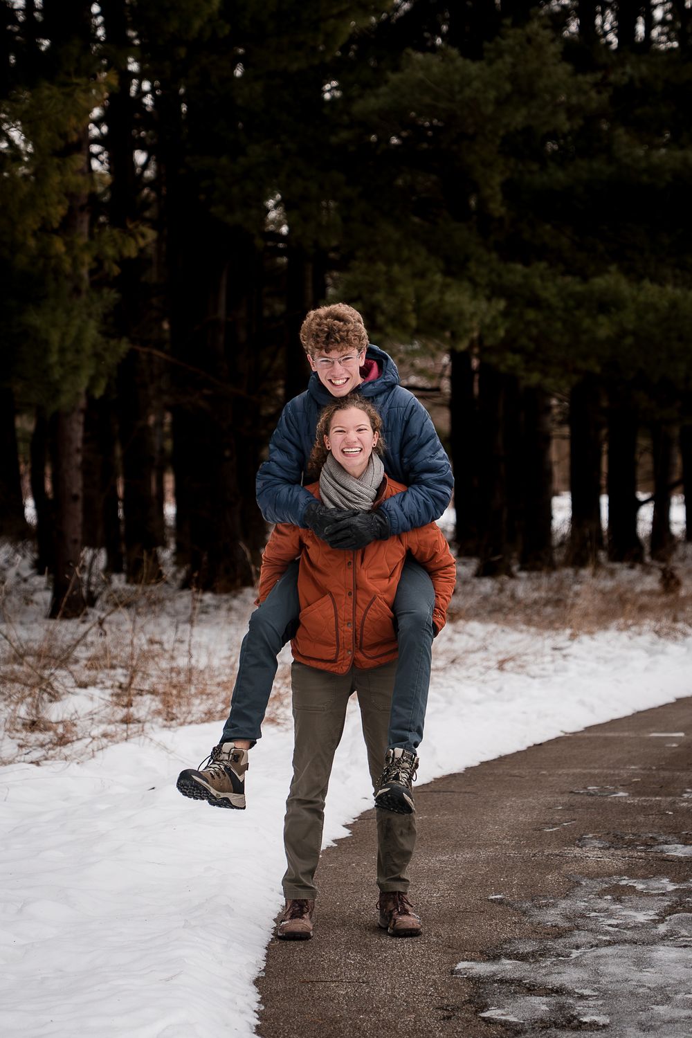 Two young people laughing on a snowy forest path, one giving the other a piggyback ride in winter coats.