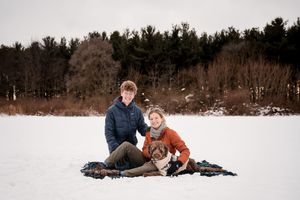 Couple sitting on blanket in snowy field with dog, evergreen trees in background, winter outdoor portrait.