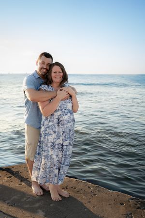 A couple embraces on a seaside pier, the woman in a blue floral dress, smiling as the ocean shimmers behind them.