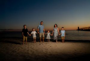 Family of six holding hands on a beach at sunset with a lighthouse in the background.