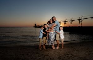 Family of five posing on sandy beach at sunset with lighthouse pier glowing in background.