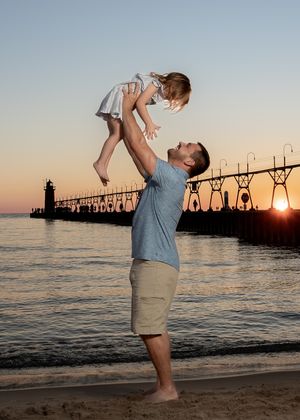 Father lifting young daughter into the air on a beach at sunset with a lighthouse pier in the background.