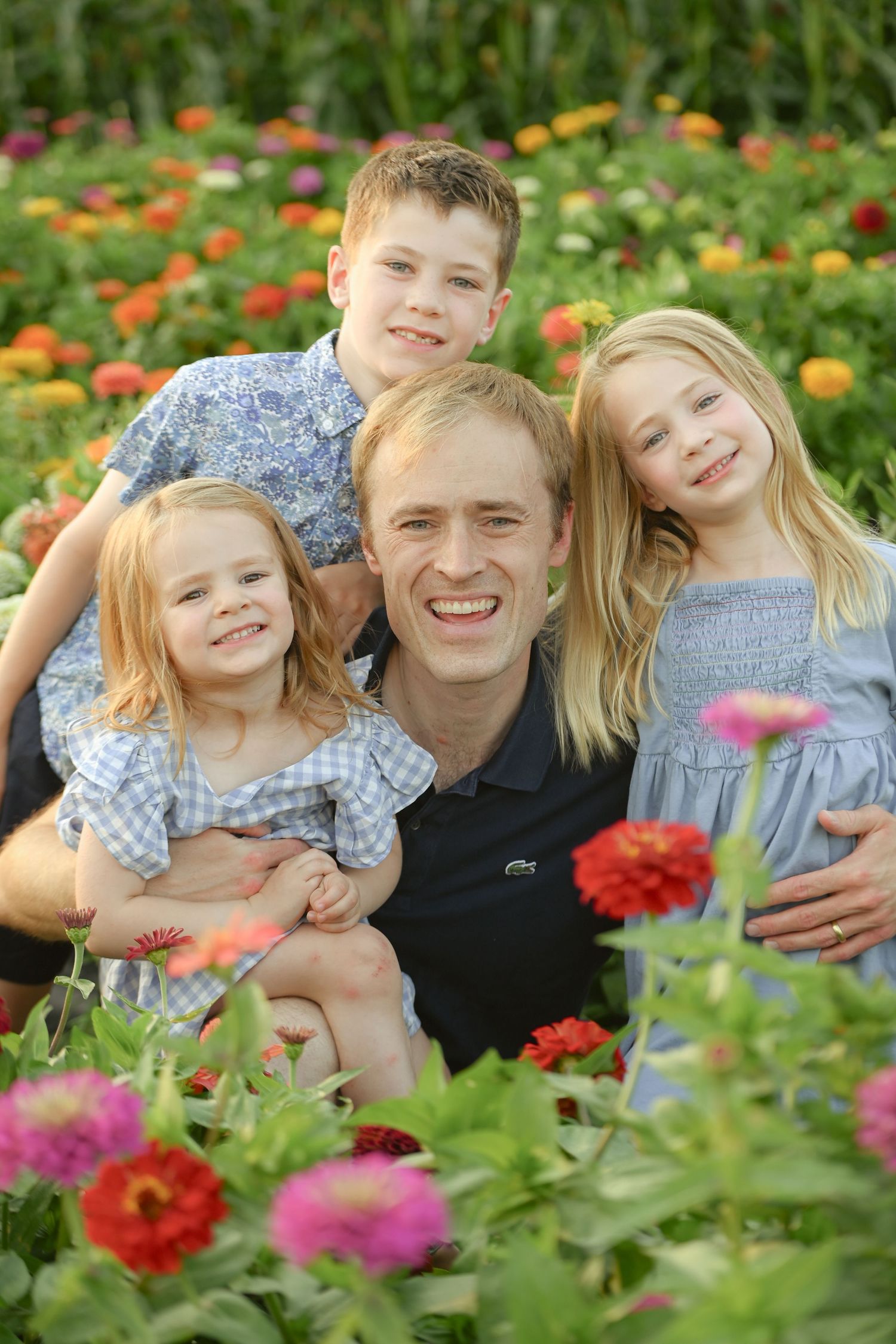 A smiling father poses with three young children in a vibrant flower field filled with colorful zinnias.