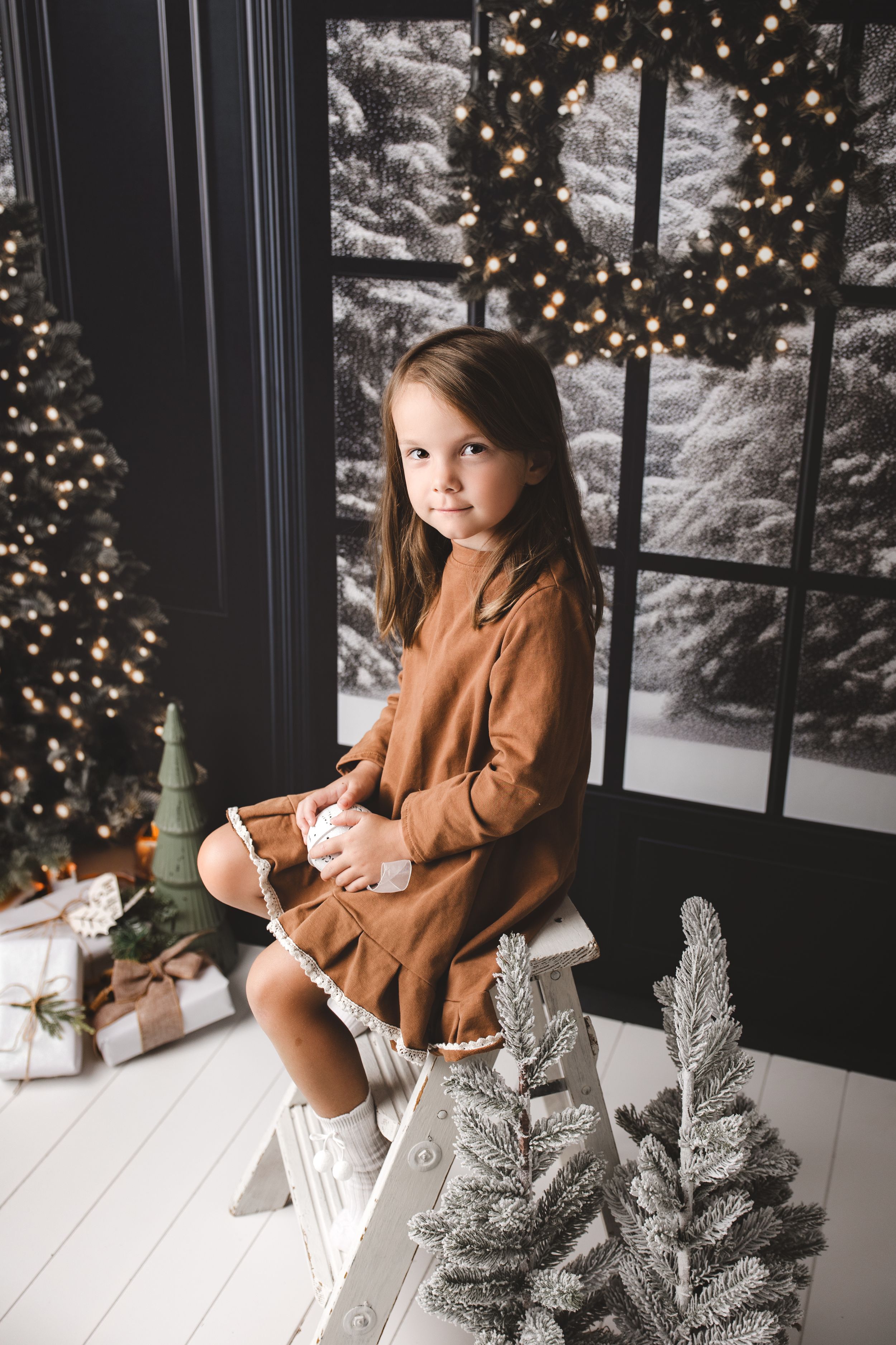 Christmas scene with a toddler girl sitting on a white step ladder. Snowy trees behind a window in the background. Presents on the flor