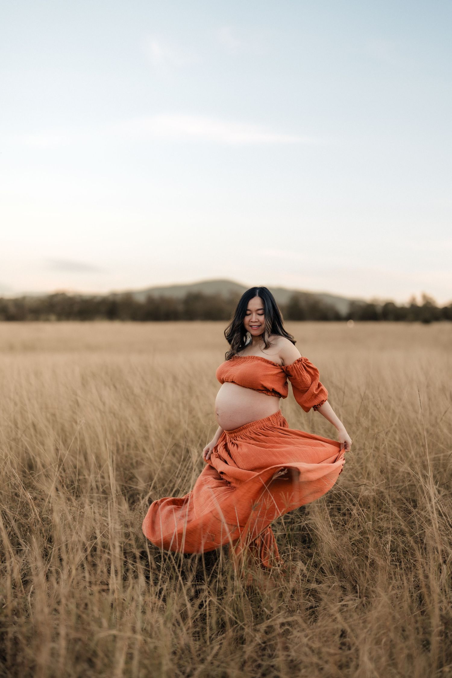 Pregnant woman in rust orange off-shoulder dress posing in golden field at sunset.