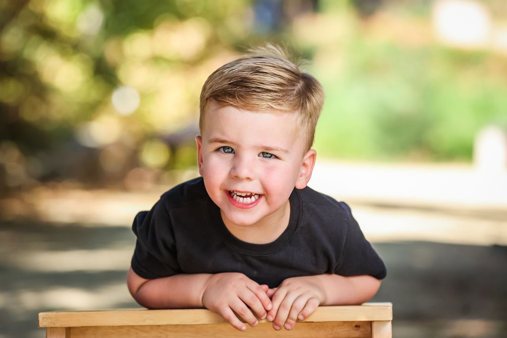 A smiling young boy with blonde hair wearing a black t-shirt, leaning on a wooden bench outdoors.