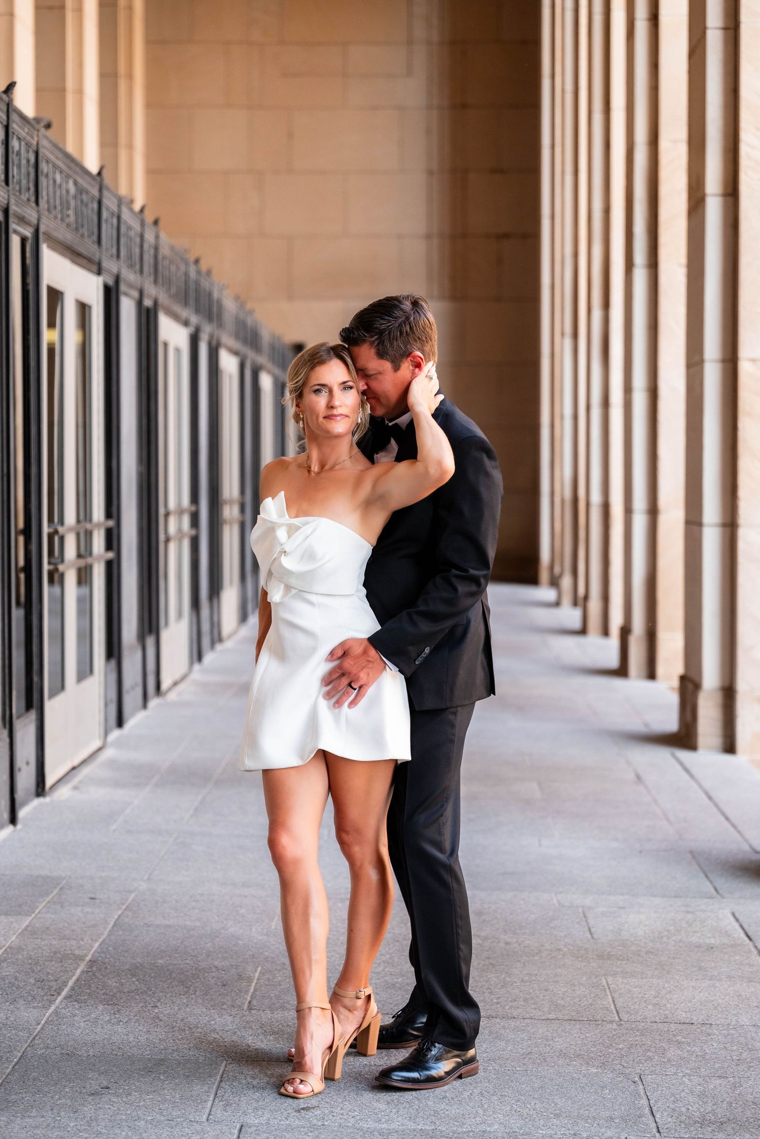 Couple in elegant attire embracing in a columned hallway, woman in white mini dress and man in dark suit.