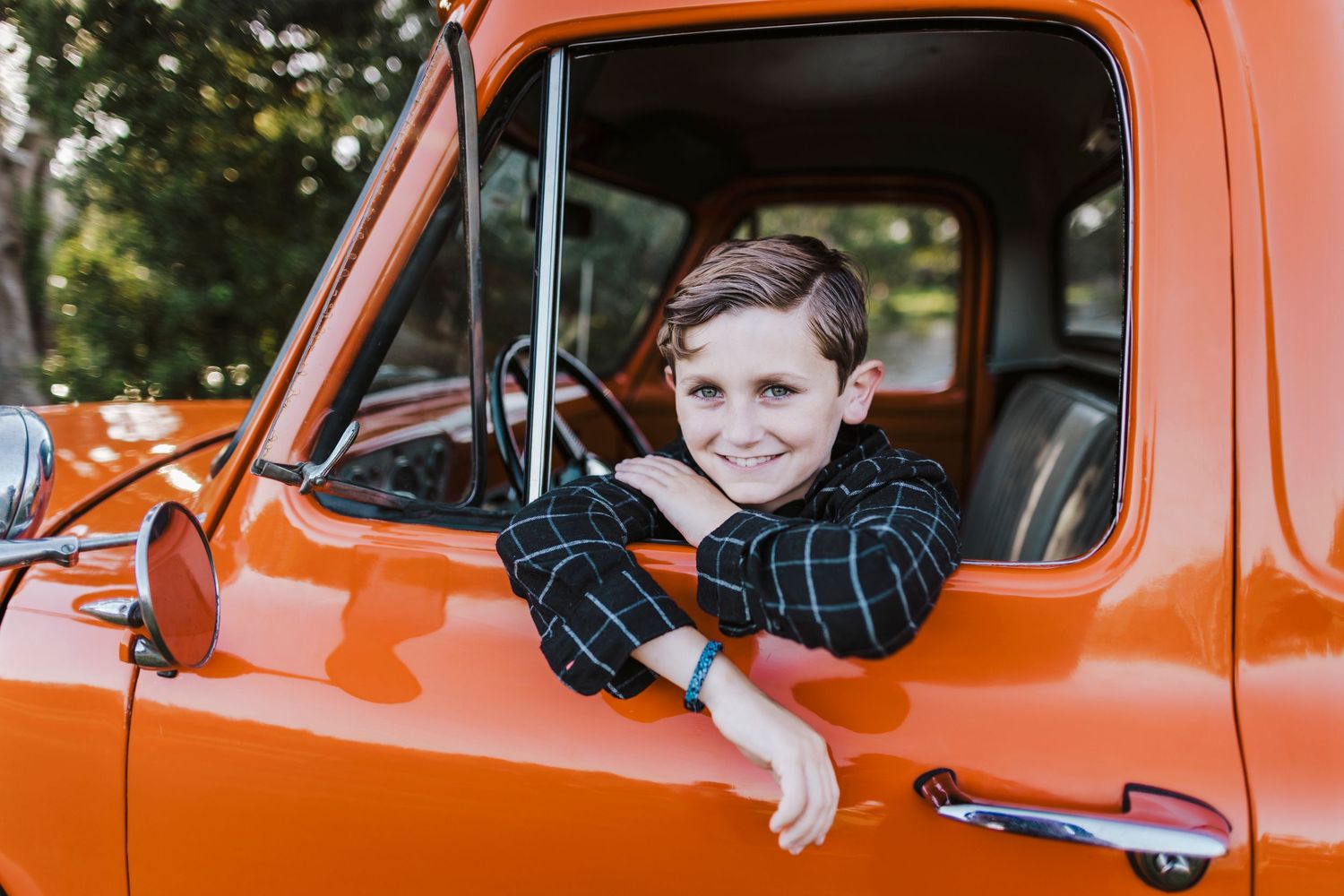 Smiling boy leaning out of a vintage orange truck window, wearing a plaid shirt and teal bracelet.