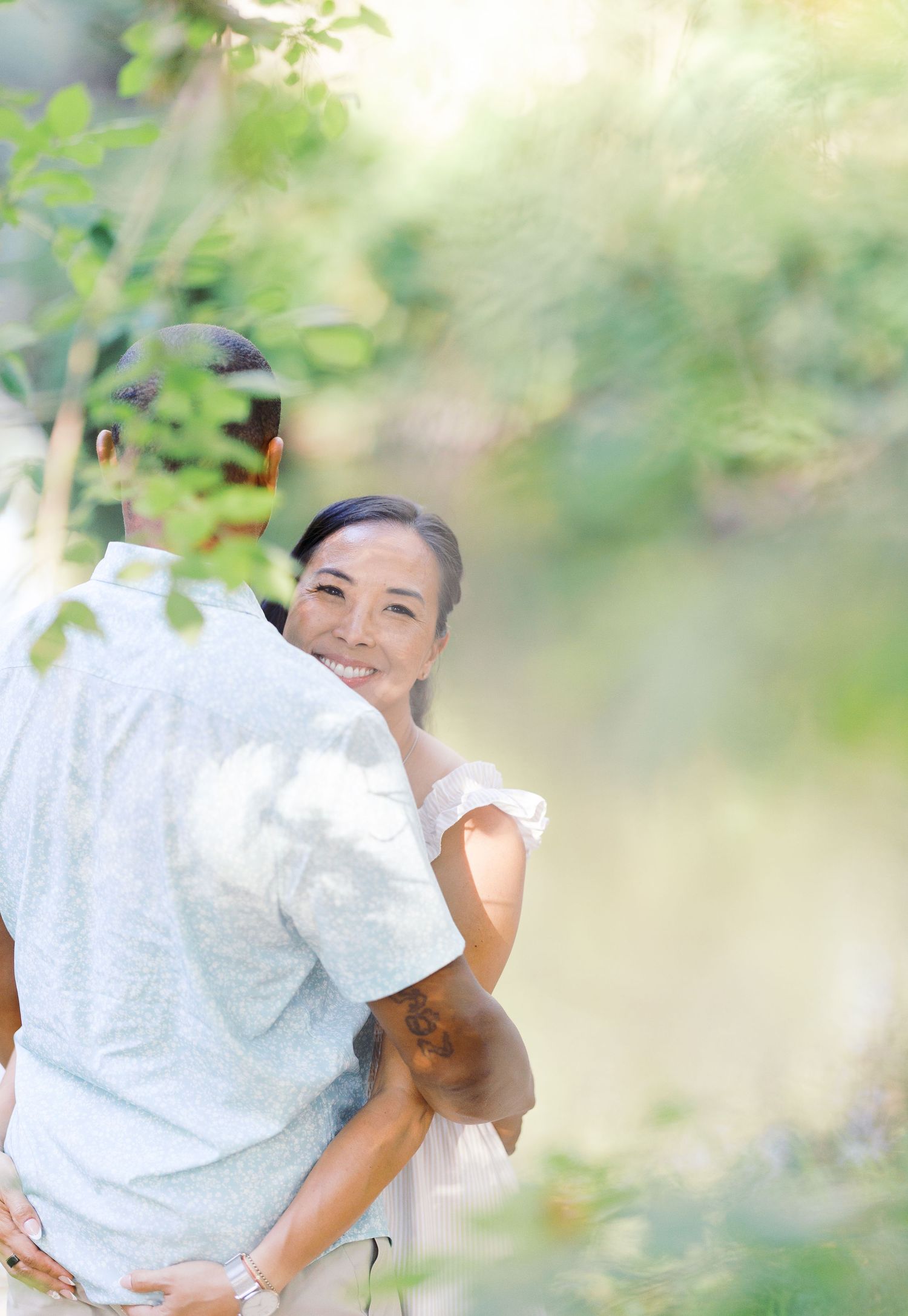 A mother in a floral dress holds her child in a sunlit green field with trees in the background.