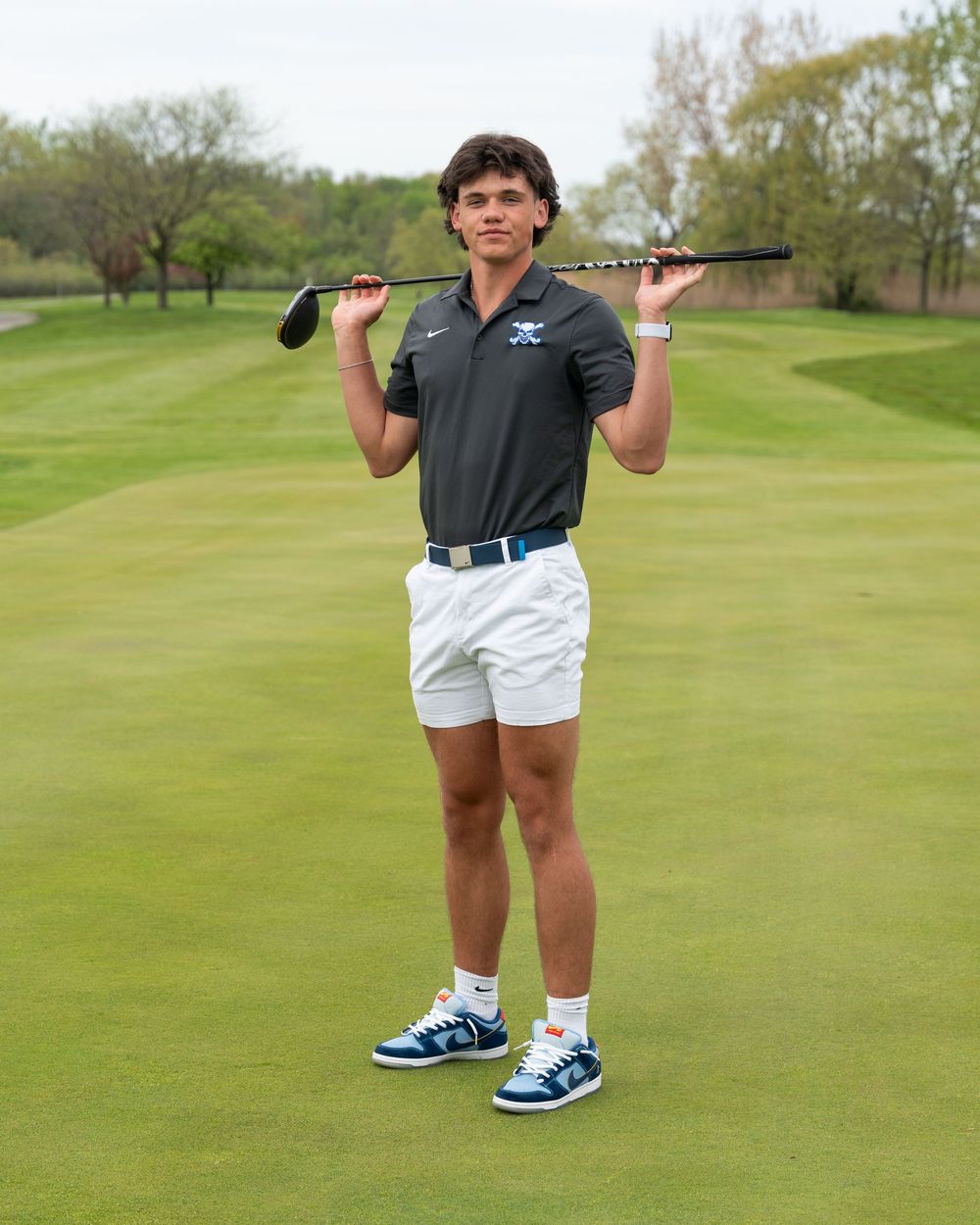 Young male golfer poses on a green fairway holding a golf club behind his shoulders wearing white shorts and a dark polo shirt.