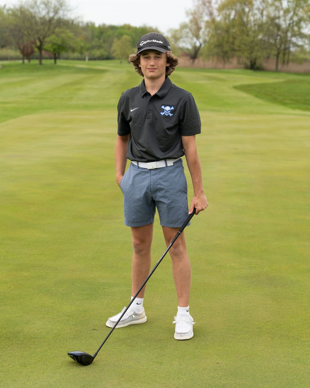 Young male golfer in black Nike polo and gray shorts poses with a driver on a lush green golf course.