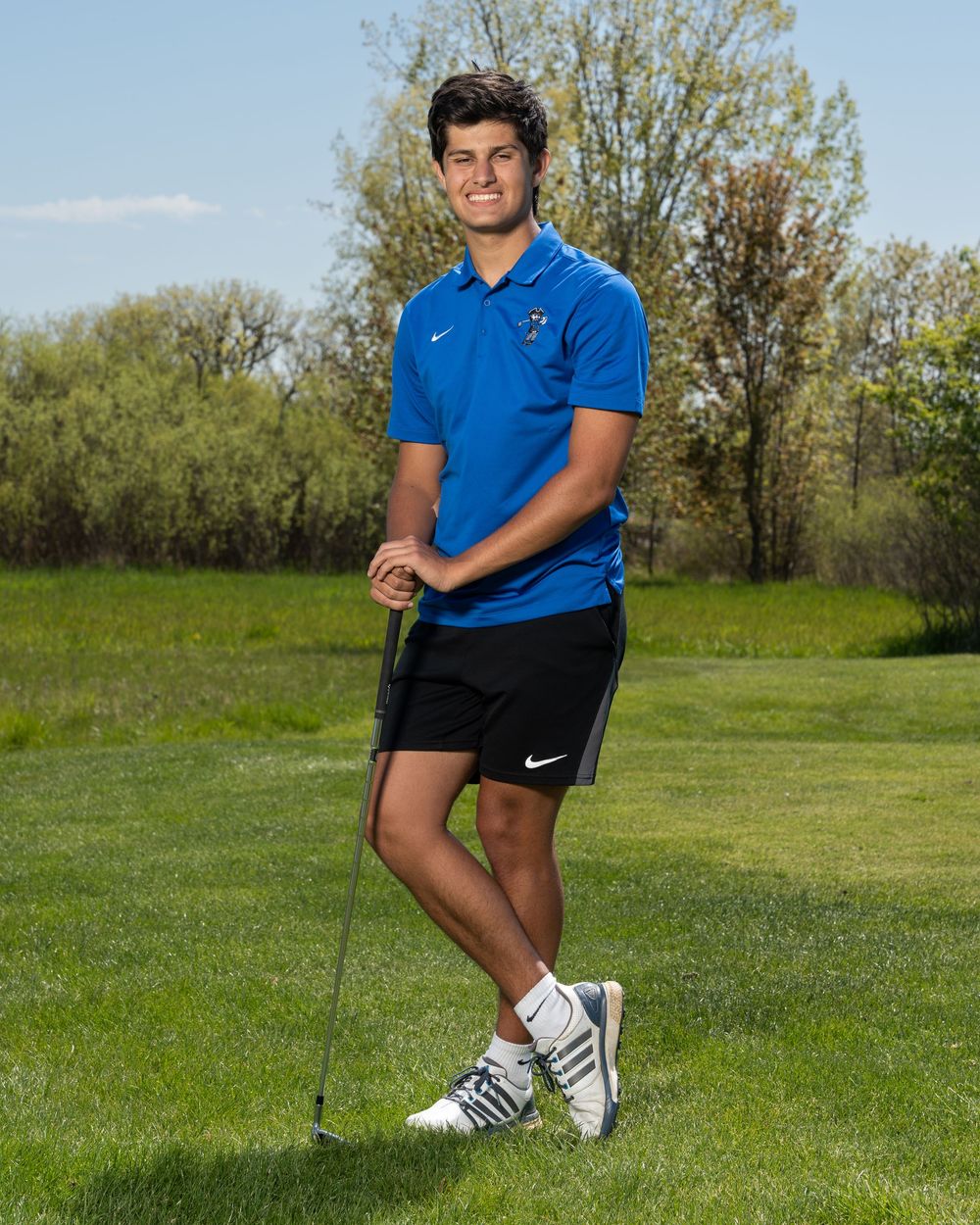 Young male golfer posing with a golf club on a green course, wearing a blue Nike polo and black shorts.