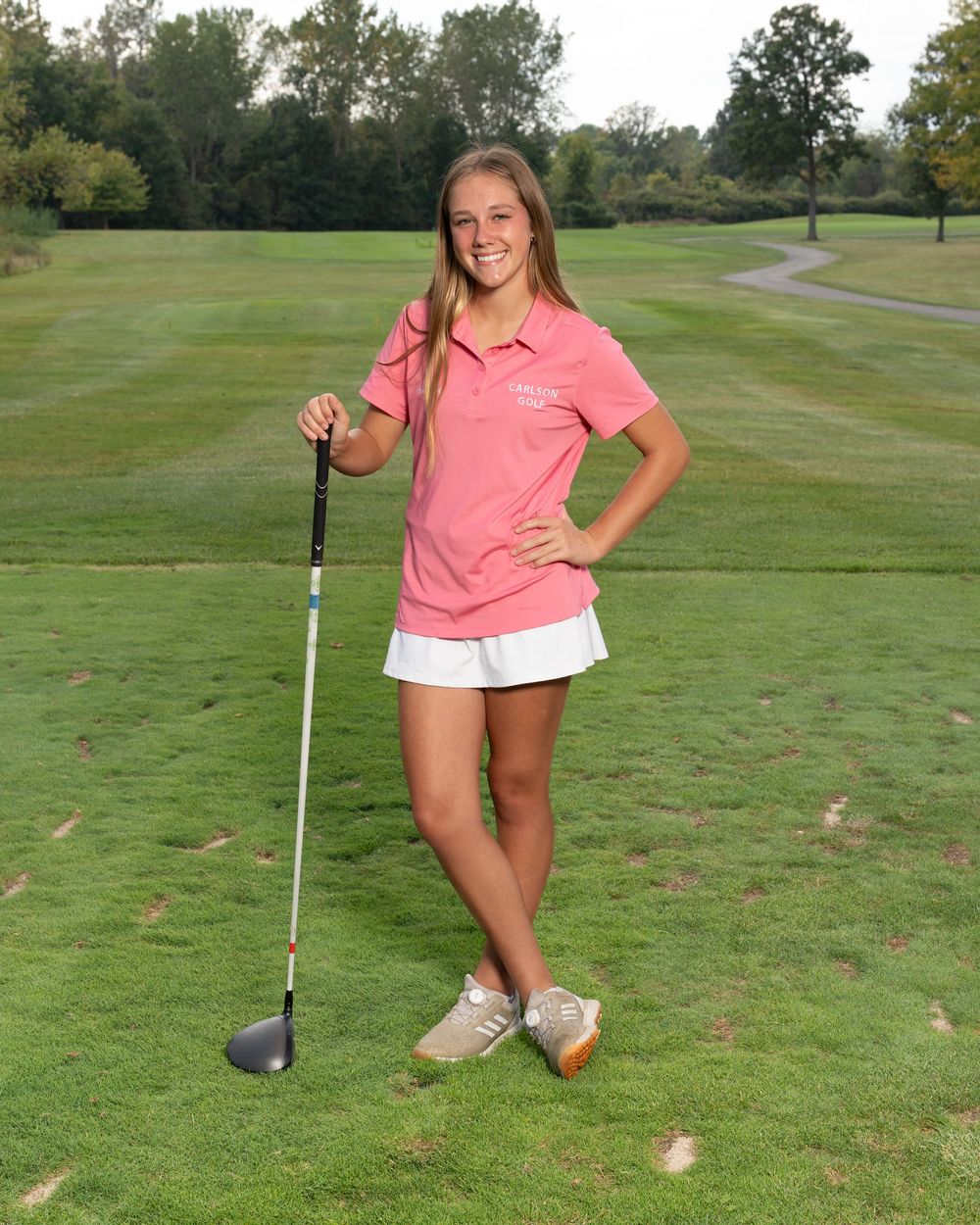Young female golfer poses on green course holding a driver club, wearing pink polo shirt and white skirt.