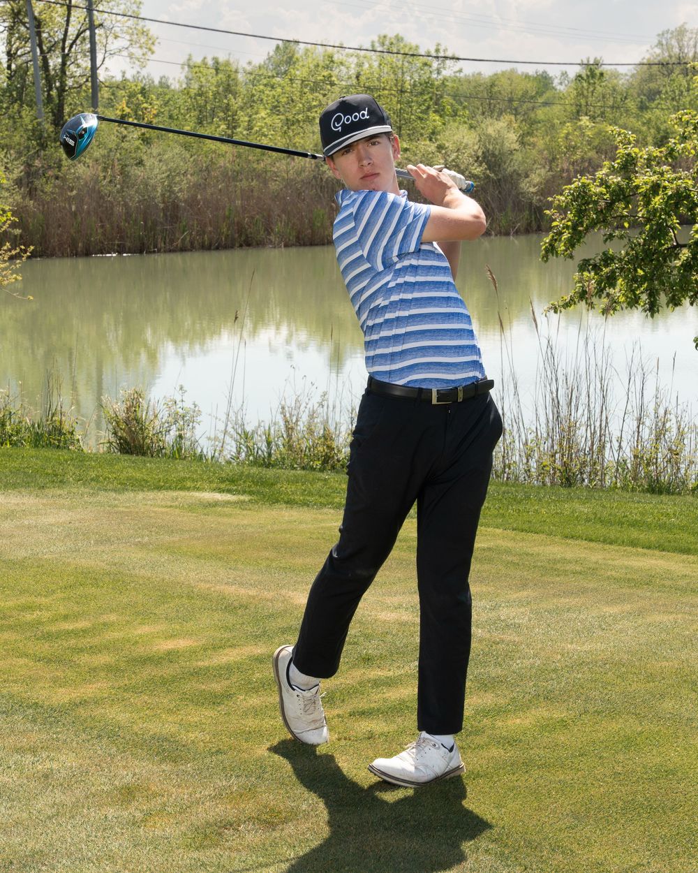 Young golfer in blue striped shirt and black pants swings a driver on a lush green course beside a serene pond.