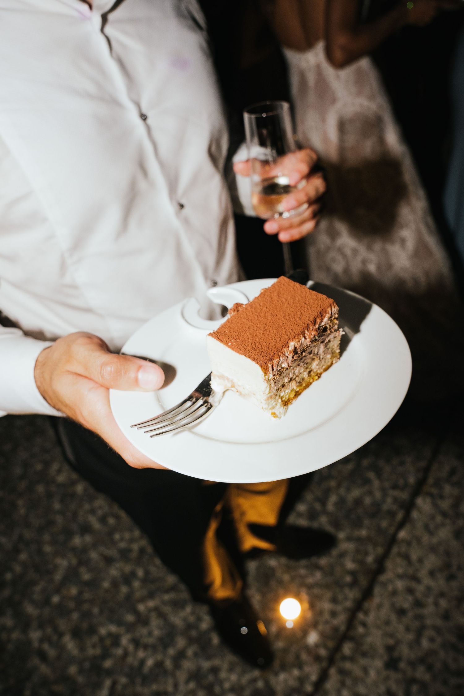 Person holding a white plate with a slice of tiramisu cake and a fork, with a champagne glass in the background.