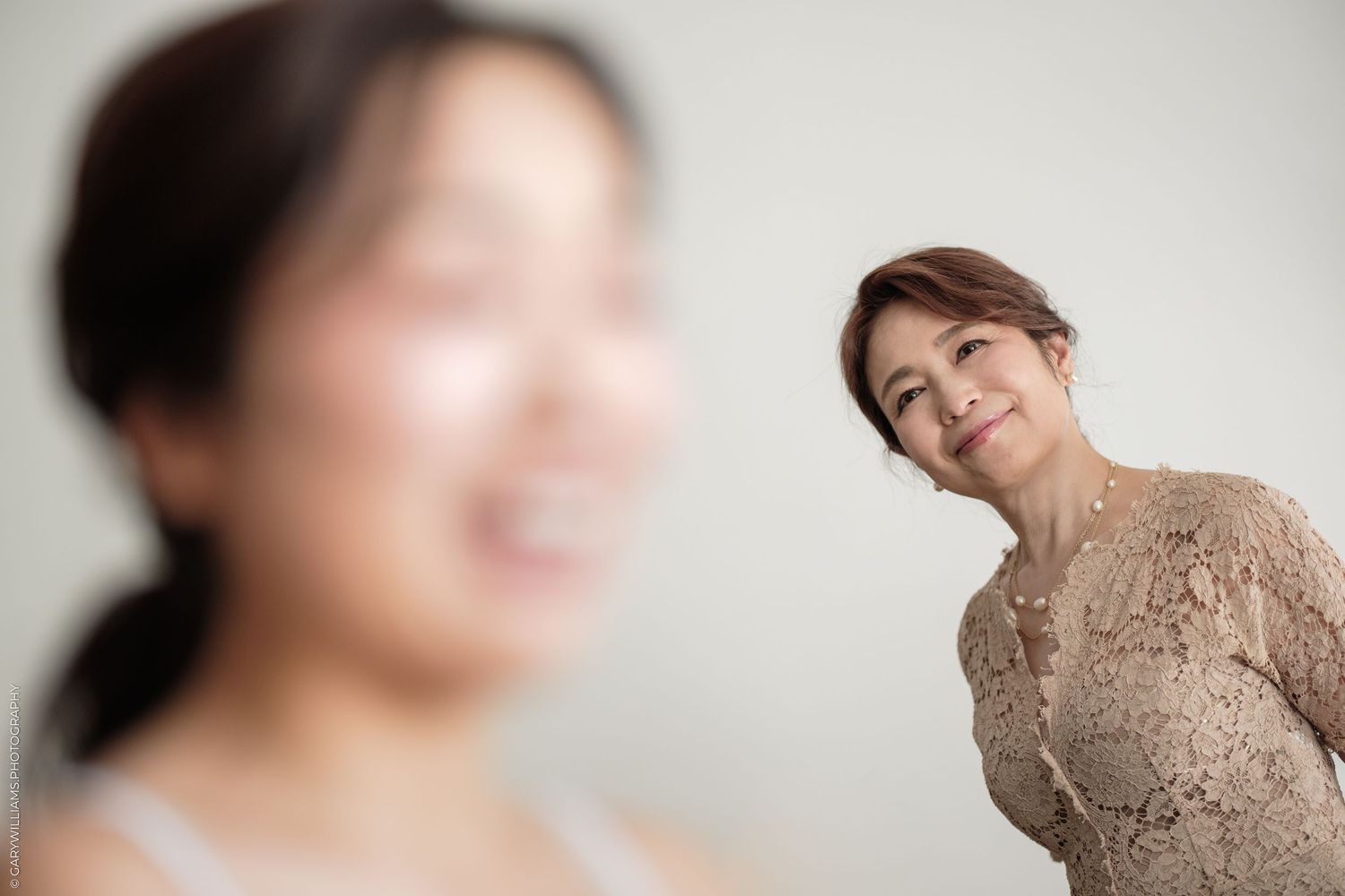 Two Asian women smiling in a studio, one blurred in foreground, older woman in lace top posing in background.