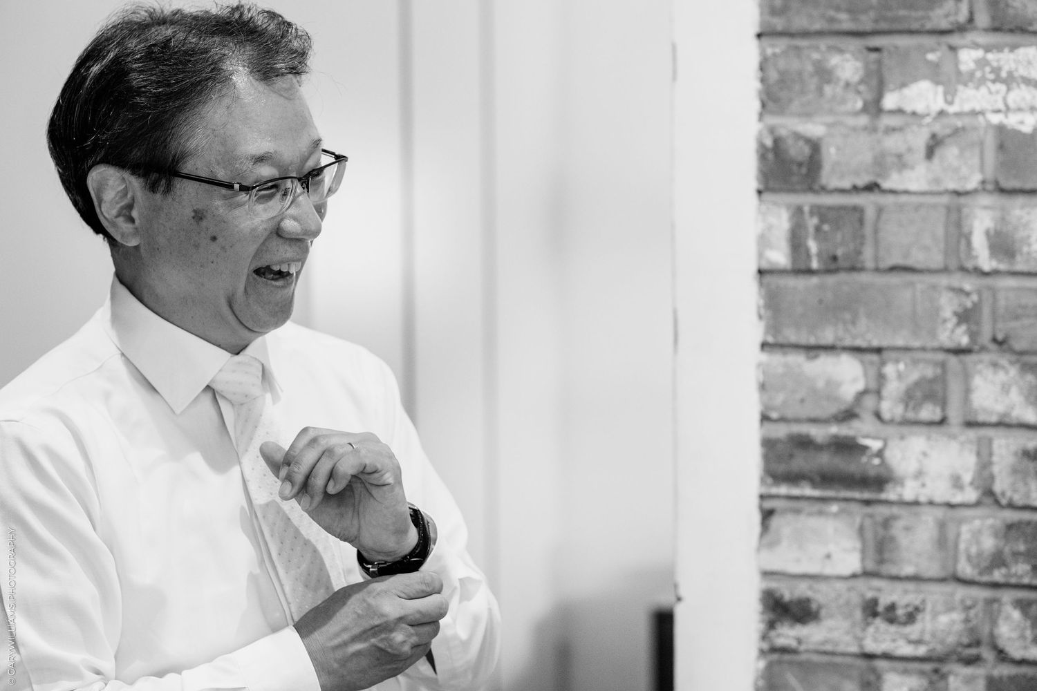 Black and white photo of a smiling man in a white dress shirt and tie adjusting his cufflinks near a brick wall.