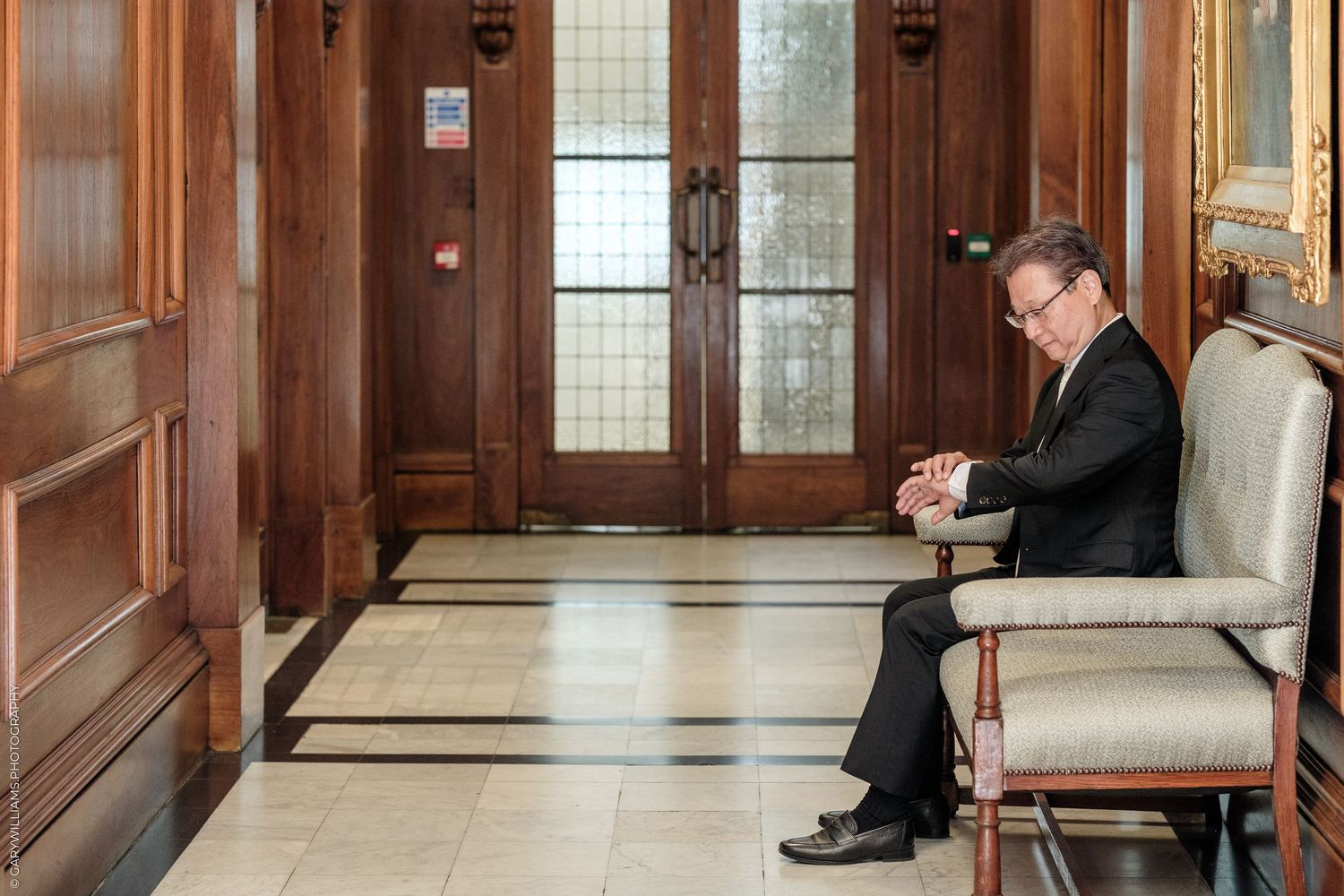 Man in black suit sitting alone in elegant wood-paneled hallway with marble floors, looking contemplative.