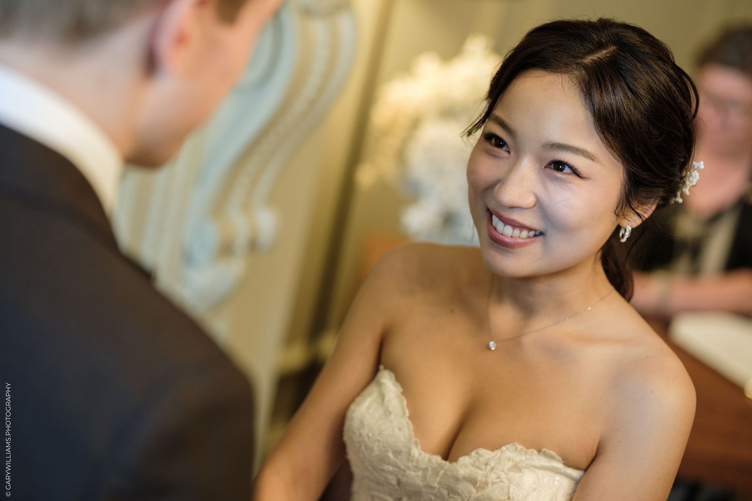 Smiling Asian bride in strapless lace wedding dress looking up at groom during wedding ceremony.