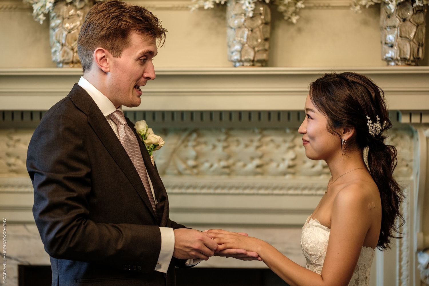 Bride and groom exchange vows holding hands at elegant wedding ceremony near ornate fireplace.