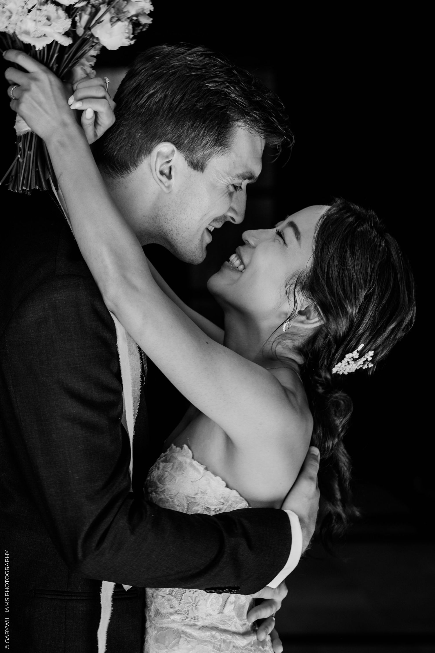 Black and white wedding photo of bride and groom embracing, laughing joyfully, bride holding bouquet aloft.