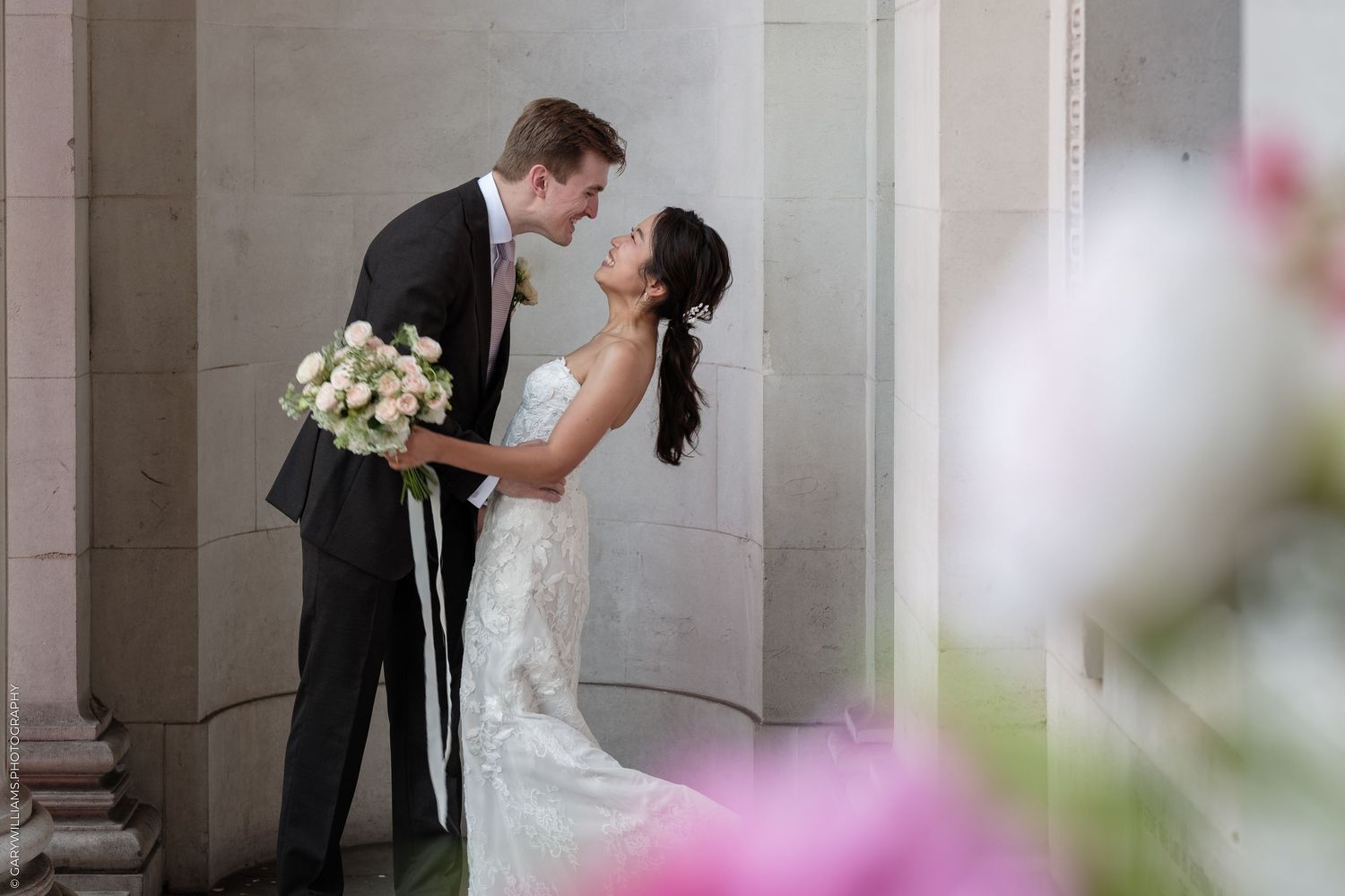 Bride and groom share a romantic moment among elegant columns, with pink flowers in the foreground.