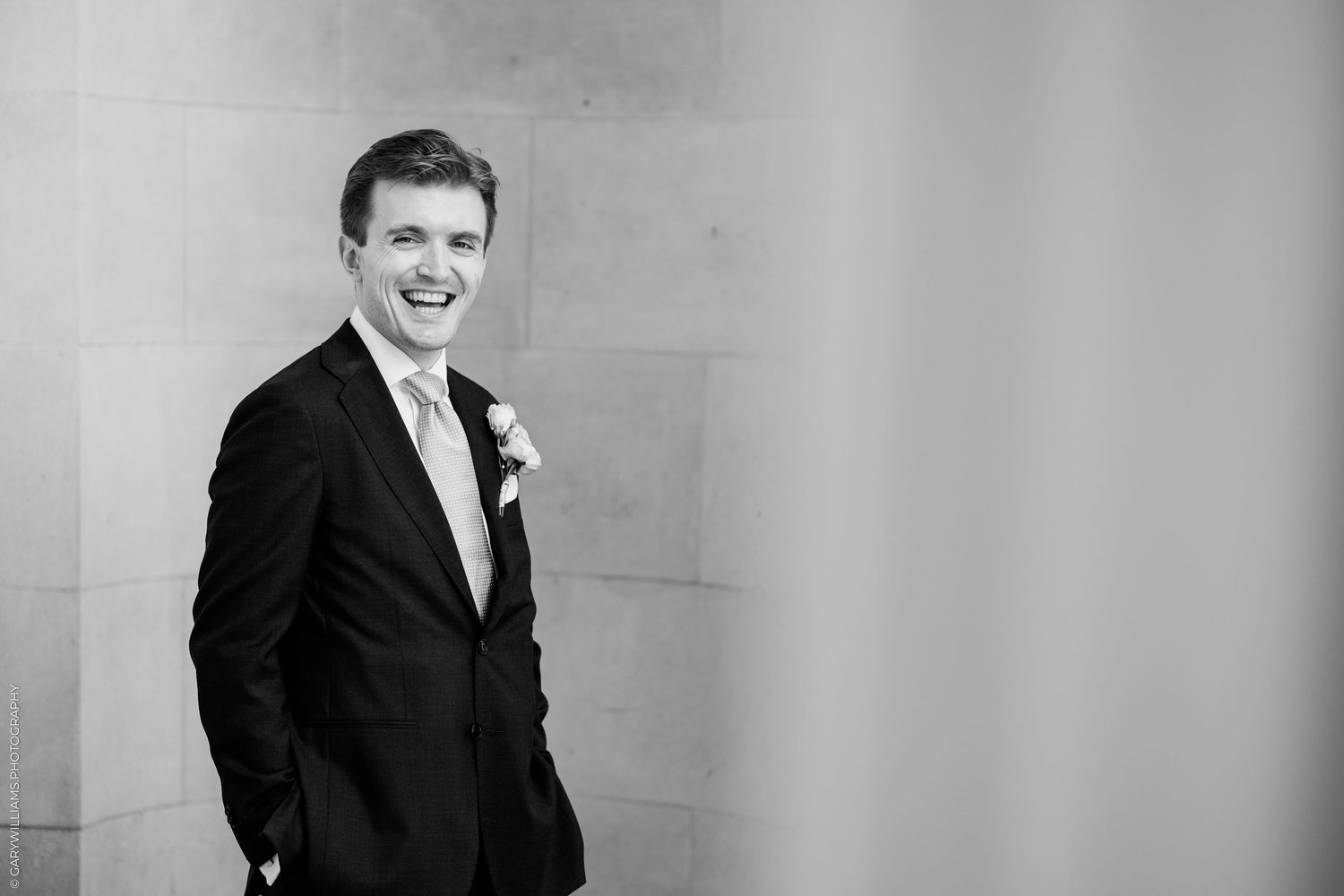 Black and white photo of a smiling groom in a dark suit with boutonniere, standing against a stone wall.