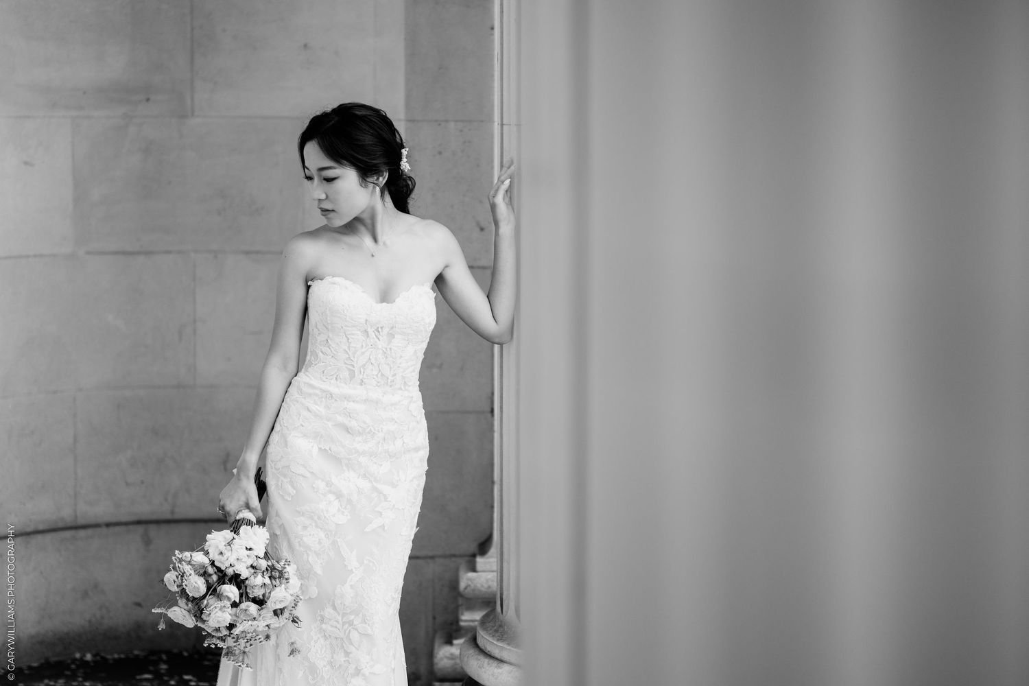 Elegant bride in a strapless lace wedding gown holding a bouquet, posing gracefully by a stone column in black and white.