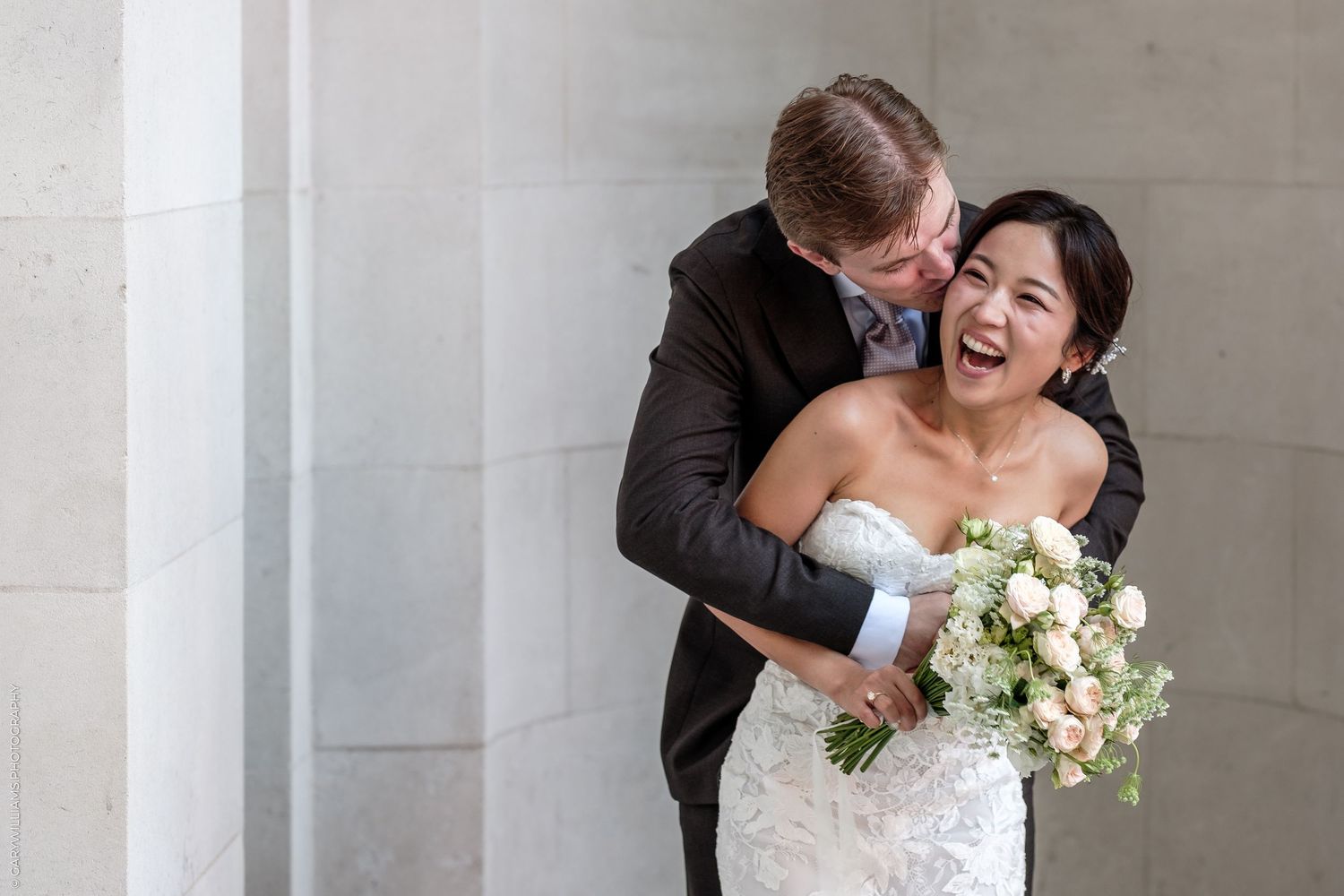 Joyful bride in lace gown holding white bouquet laughs as groom in gray suit embraces her near stone columns.
