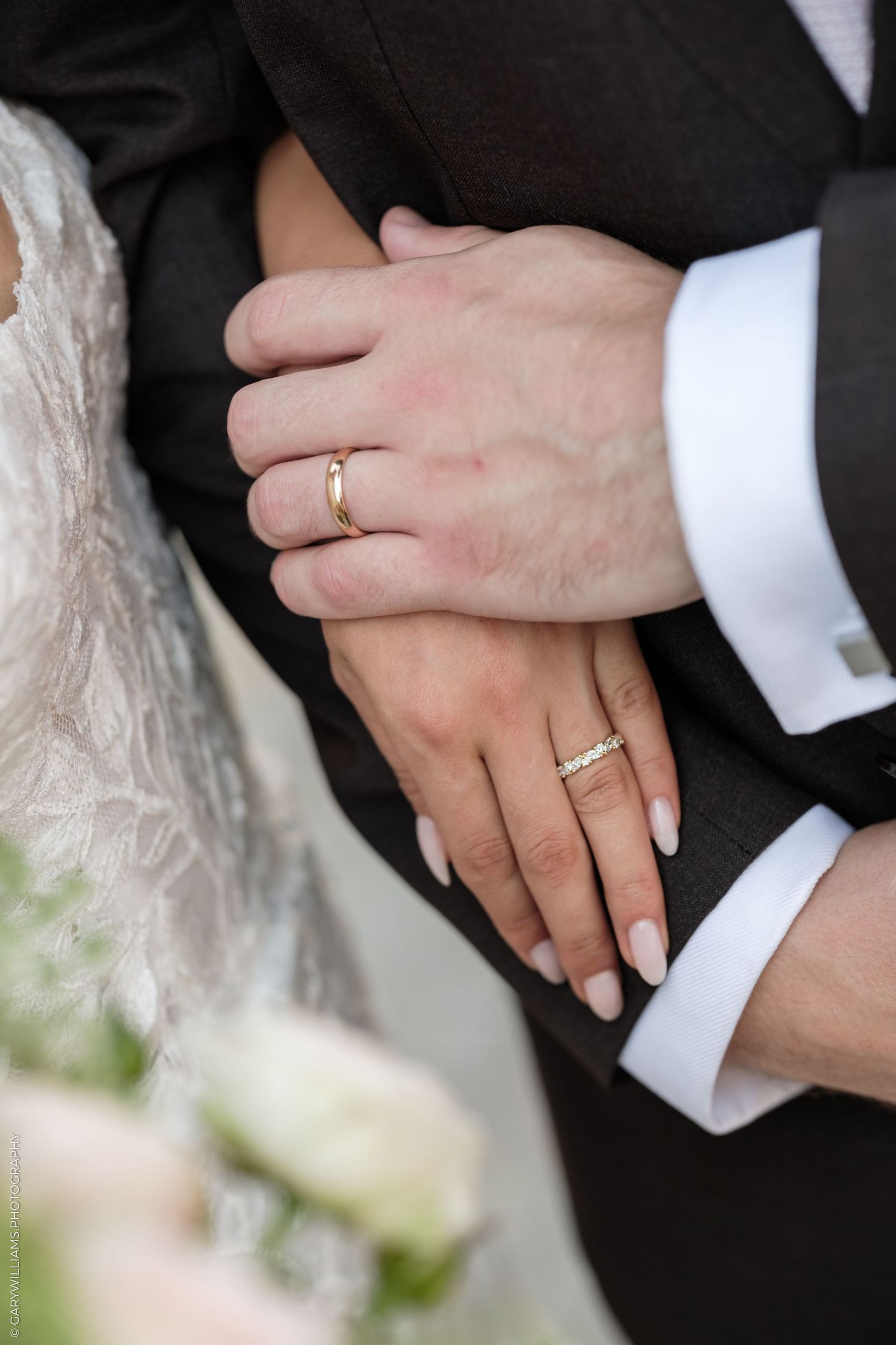 Newlyweds displaying gold wedding rings on their hands, bride in lace dress holding grooms arm in black suit.