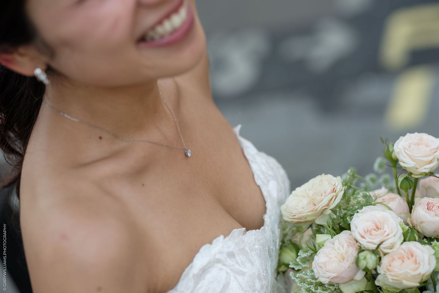 Smiling bride in white lace dress holding a bouquet of soft pink roses, wearing a delicate silver necklace.