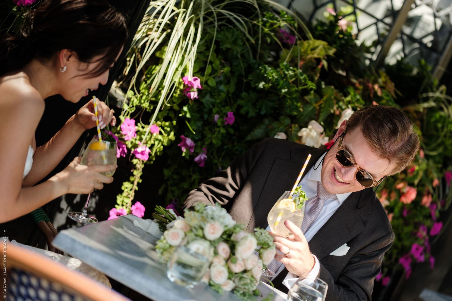 Couple enjoying cocktails at an outdoor garden restaurant surrounded by lush greenery and colorful flowers.
