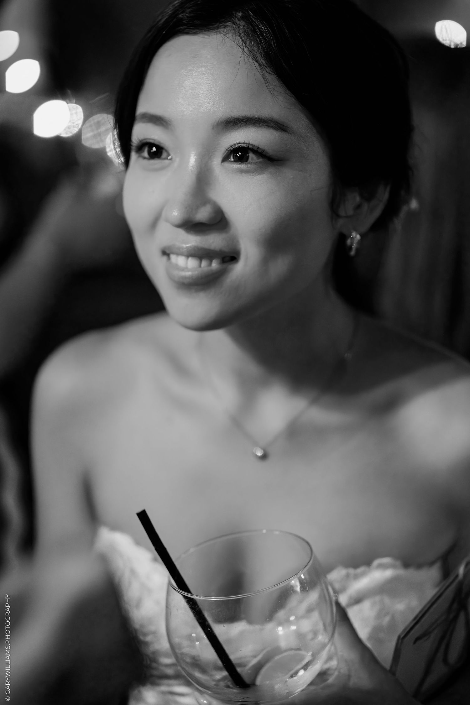 Black and white portrait of a smiling young woman holding a cocktail glass with a straw at a formal event.