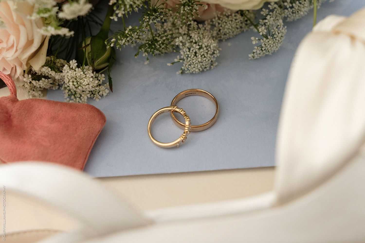 Two gold wedding bands resting on a light blue surface beside a bridal bouquet with white flowers and ribbon.