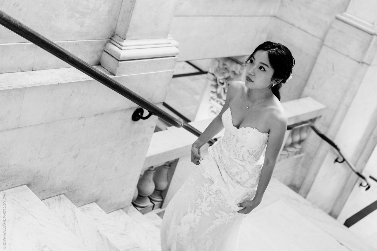 Black and white photo of a bride in a strapless lace gown descending marble stairs, holding the railing.