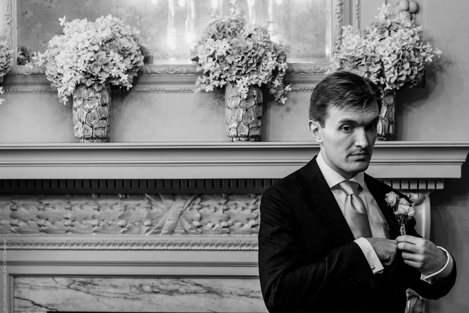 Black and white photo of a groom in a suit adjusting his cufflinks near a floral mantelpiece at a wedding.