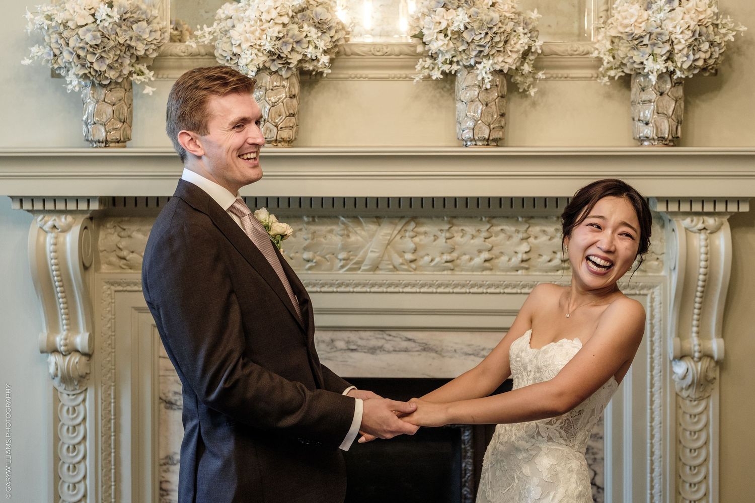 Joyful newlywed couple holding hands and laughing in front of an elegant white fireplace with floral arrangements.