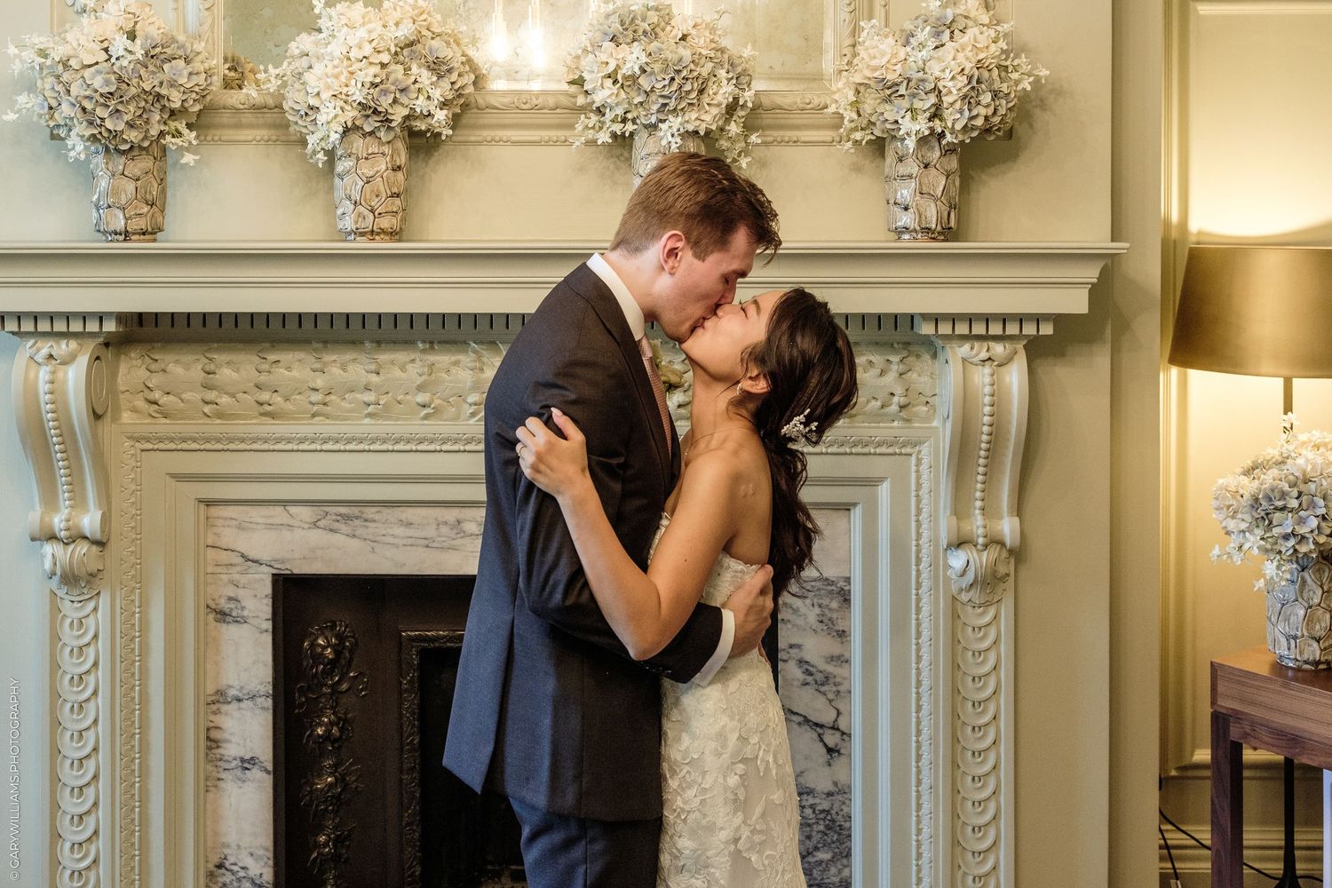 Couple sharing first kiss as newlyweds in front of elegant white marble fireplace with floral arrangements at wedding reception.