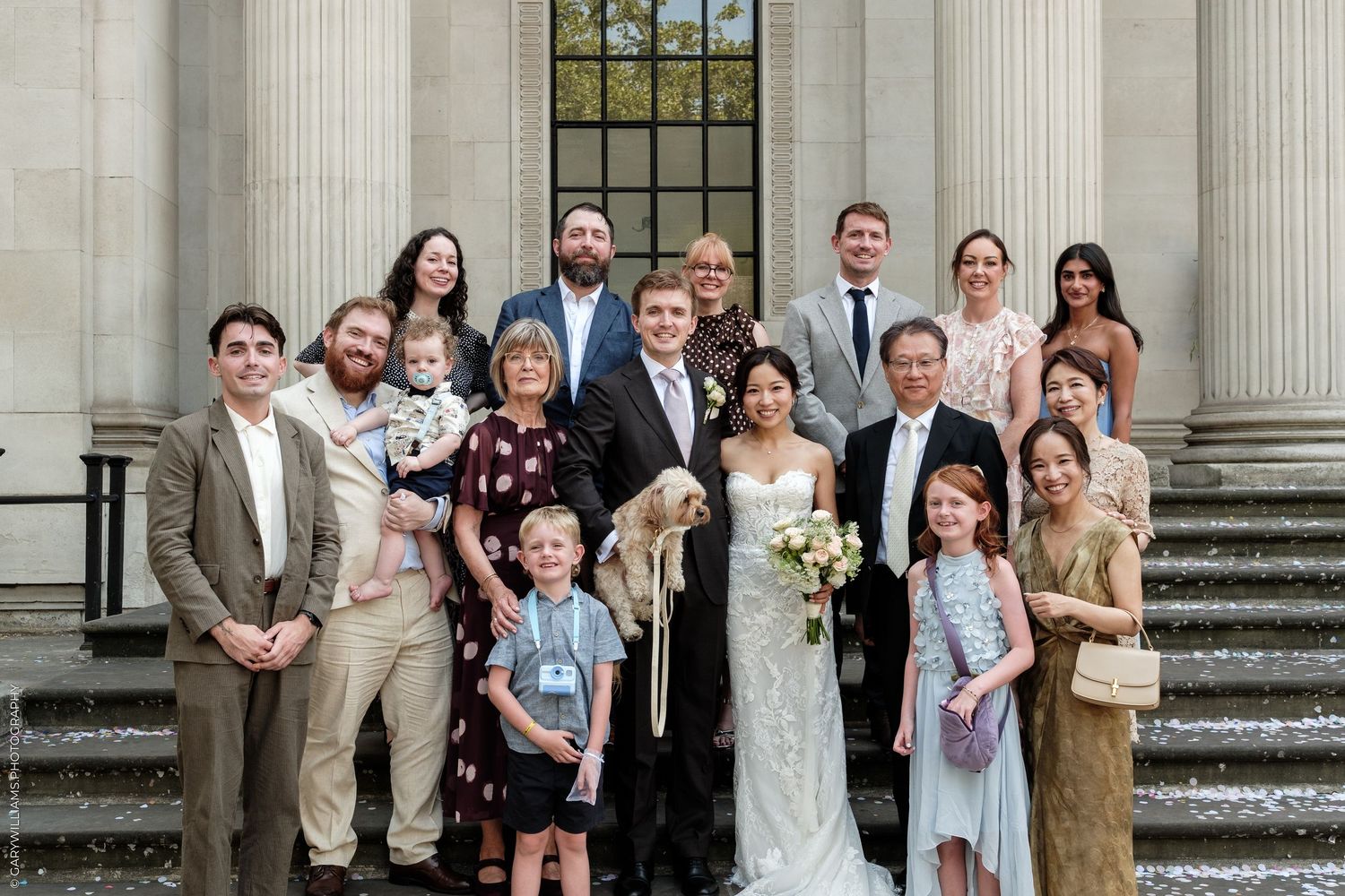 A large wedding party poses on stone steps outside a grand building, featuring the bride in white and diverse family members.