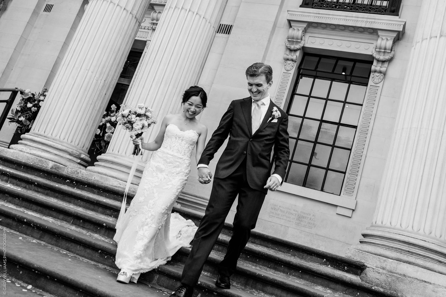 Bride and groom holding hands walking down steps of elegant columned building on their wedding day.