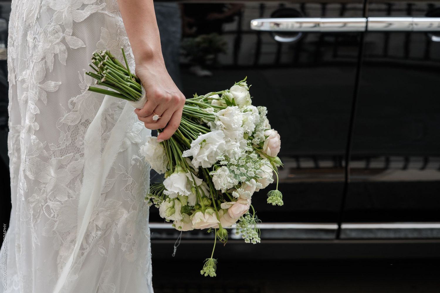 Bride holding a lush white wedding bouquet with ranunculus and greenery, wearing a lace gown near a black vehicle.