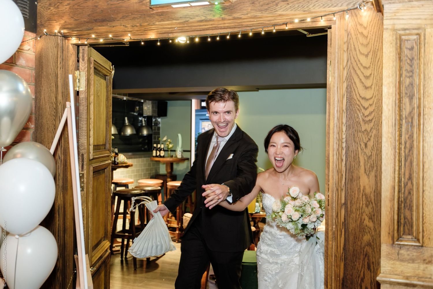 Joyful bride and groom making grand entrance at wedding reception, bride holding white bouquet, balloons in background.