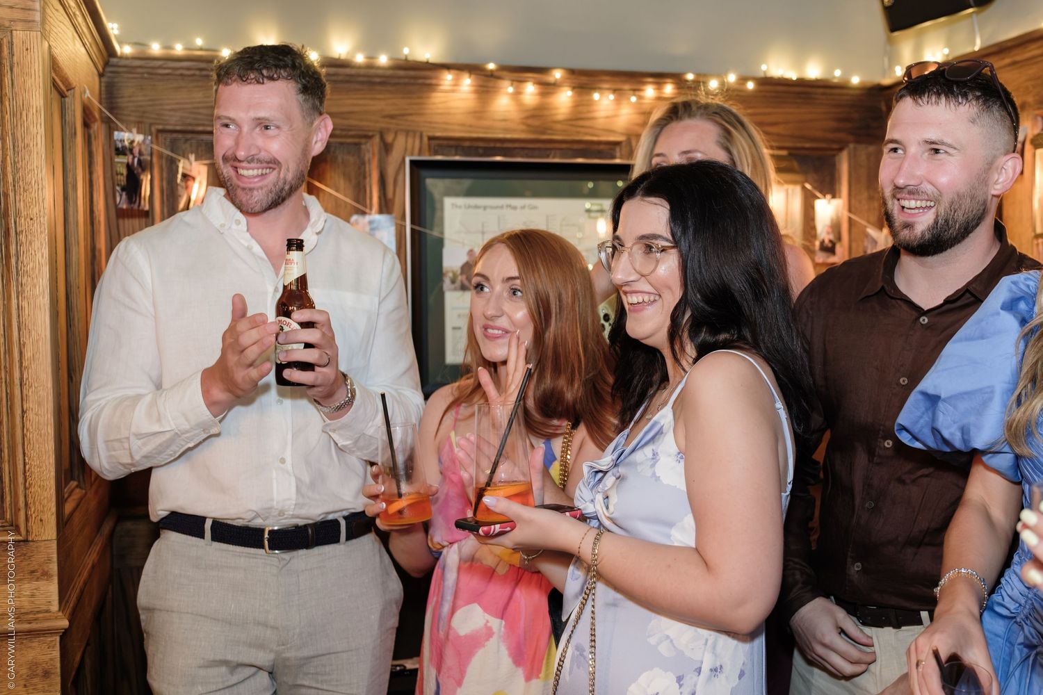 Group of friends laughing and socializing at an indoor party, holding drinks in a warmly lit venue.