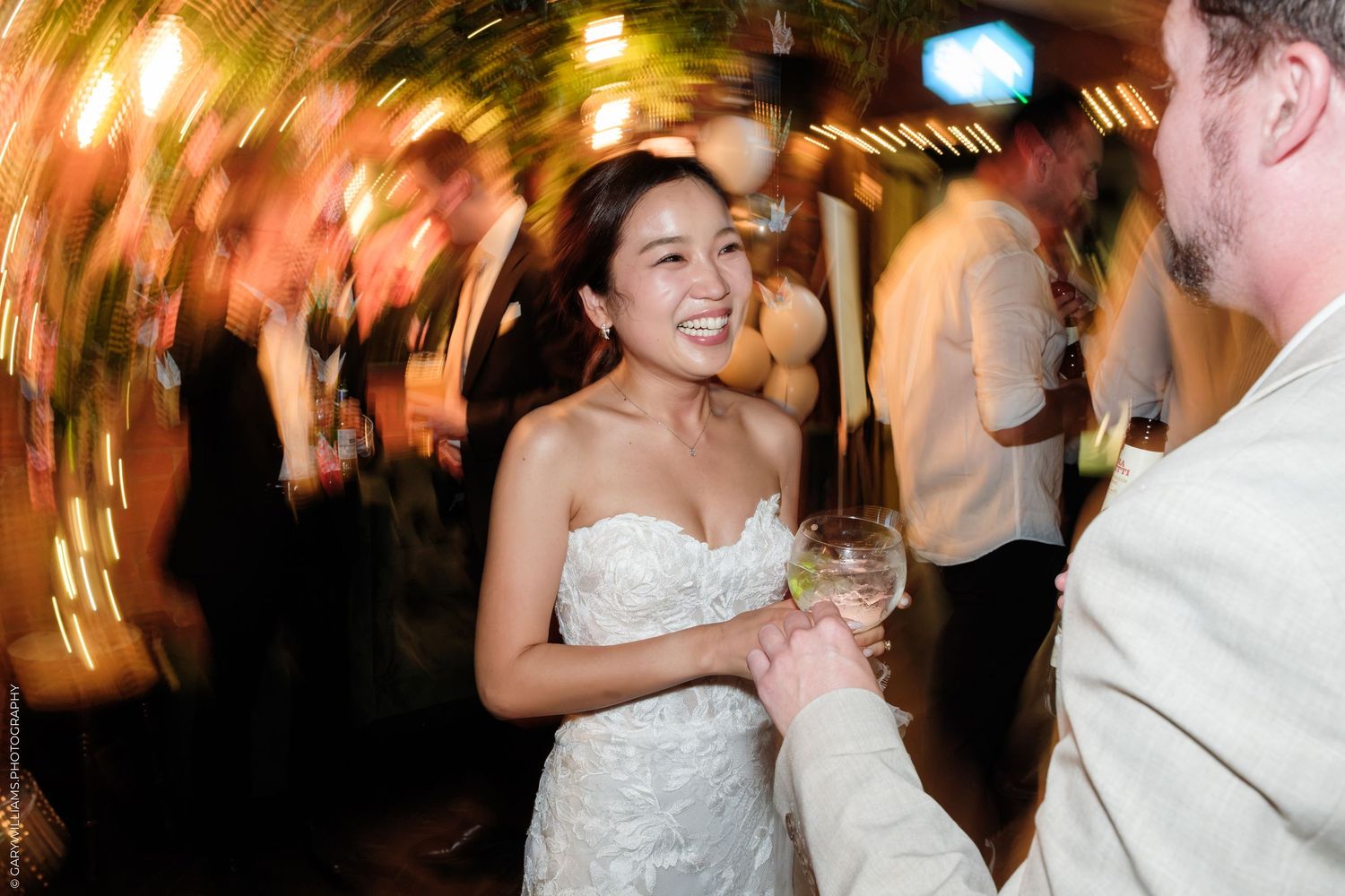 Bride in strapless lace gown smiling and holding wine glass with groom at lively wedding reception.